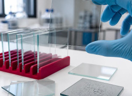 Gloved hand holding a glass test panel with water droplets, beside panels in a rack