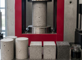 Concrete test cylinders and cubes beside a compression testing machine, with a jar of black graphene concentrate