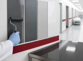 Gloved hand rolling a gray coating onto a wall test panel in a clean hospital corridor